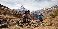 Two mountain bikers ride through an alpine landscape with the Matterhorn in the background.