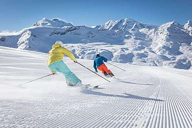 Zwei Skifahrer fahren auf einer präparierten Piste in den Bergen.