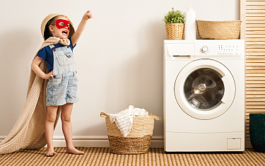 Un enfant avec un masque rouge et une cape se tient à côté d'une machine à laver.