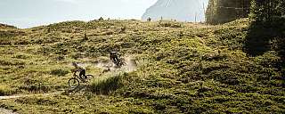 Zwei Mountainbiker fahren auf einem Pfad mit Berg im Hintergrund.