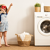 Ein Kind mit roter Maske und Umhang steht neben einer Waschmaschine.