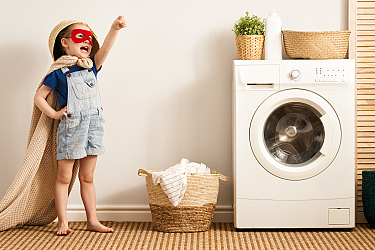 A child in a red mask and cape stands next to a washing machine.